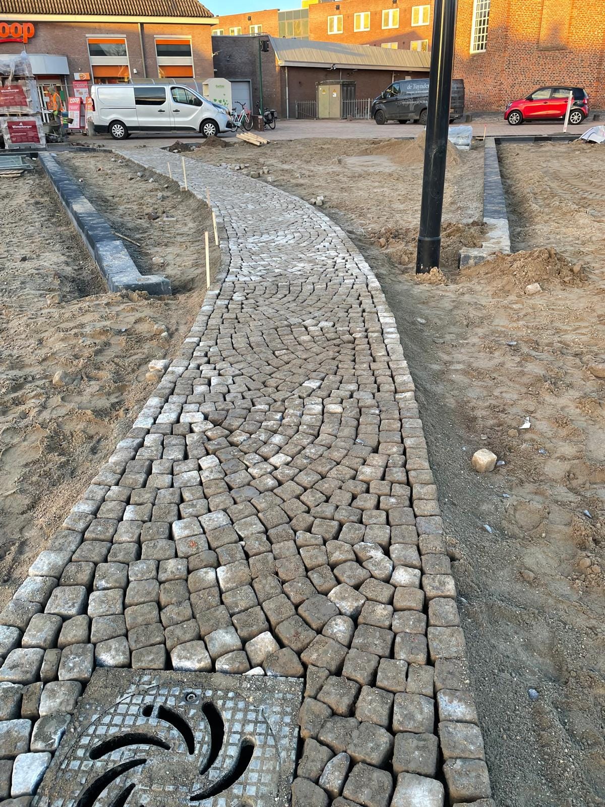 Pad van kasseien, ofwel kinderkopjes, dat leidt naar een winkelstraat bij een supermarkt, aangelegd door De Tuynderie.